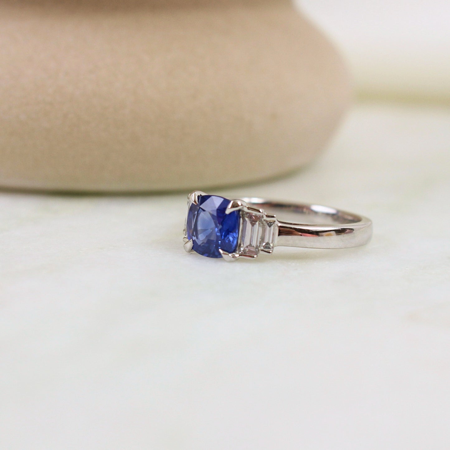 Bague de fiancaille saphir coussin épaulé par des diamants baguette en escalier sur les épaules de la pierre centrale. La bague est en or blanc 18k